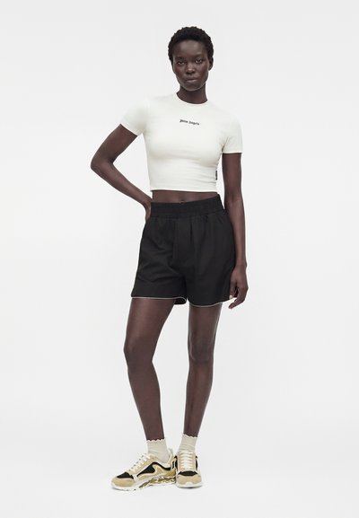 Γυναίκα όρθια με το χέρι στη μέση, φορά λευκό crop top, μαύρο σορτς, λευκές κάλτσες και μπεζ-μαύρα αθλητικά παπούτσια σε λευκό φόντο.