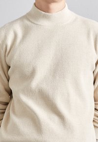 Person in einem cremefarbenen gerippten Rollkragenpullover, von den Schultern bis zur Mitte des Oberkörpers zu sehen, vor einem schlichten Hintergrund.