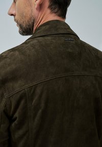 Homme avec des cheveux courts et une barbe portant une veste en suède olive foncé, montré de dos et en profil latéral contre un fond uni.