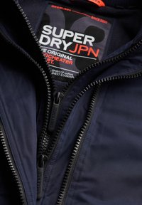 Gros plan sur une veste marine avec deux fermetures éclair noires et une étiquette de marque Superdry JPN visible à l'intérieur du col.