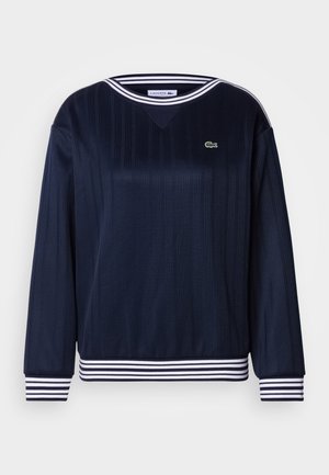 Tamsiai mėlynas ilgomis rankovėmis džemperis su ribsine faktūra, baltais dryžuotais rankogaliais ir juosmeniu, V formos iškirpte ir mažu žaliu krokodilo logotipu.