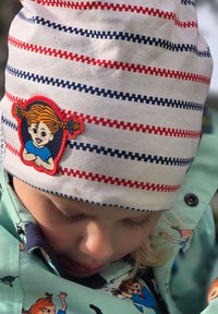 Raidallista kankaat beanie valkoisena, punaisena ja tummansinisenä, jossa on brodeerattu tyttöhahmo. Lapsen piparmintunvihreässä takissa on leikkisiä hahmosuunnitteluja.