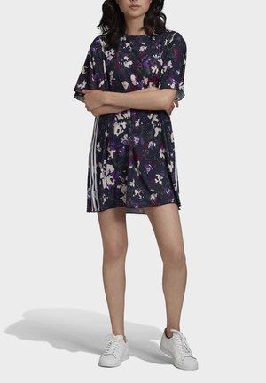 Femme portant une robe courte à manches courtes à fleurs foncées avec des rayures blanches sur les côtés, debout les bras croisés, chaussée de baskets blanches.