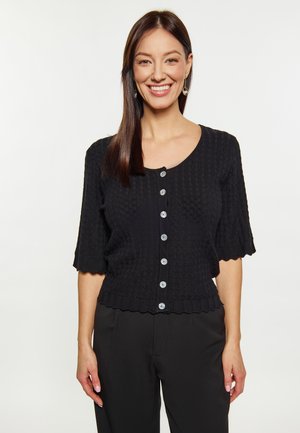Schwarze gestrickte Strickjacke mit einem strukturierten Muster, kurzen Ärmeln und gewelltem Saum. Verfügt über fünf hellfarbige Knöpfe auf der Vorderseite.