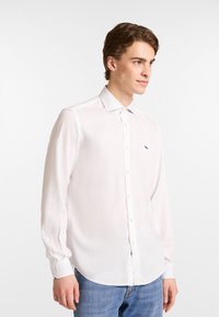 Witte blouse met lange mouwen, een klassieke kraag, knoopsluiting en een klein marineblauw logo op de borst, gemaakt van een gladde, lichte stof.