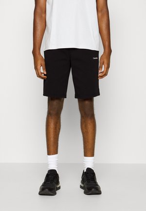 Homme portant un short Calvin Klein noir, des chaussettes blanches montantes, des baskets noires et un t-shirt blanc, debout devant un fond clair uni.