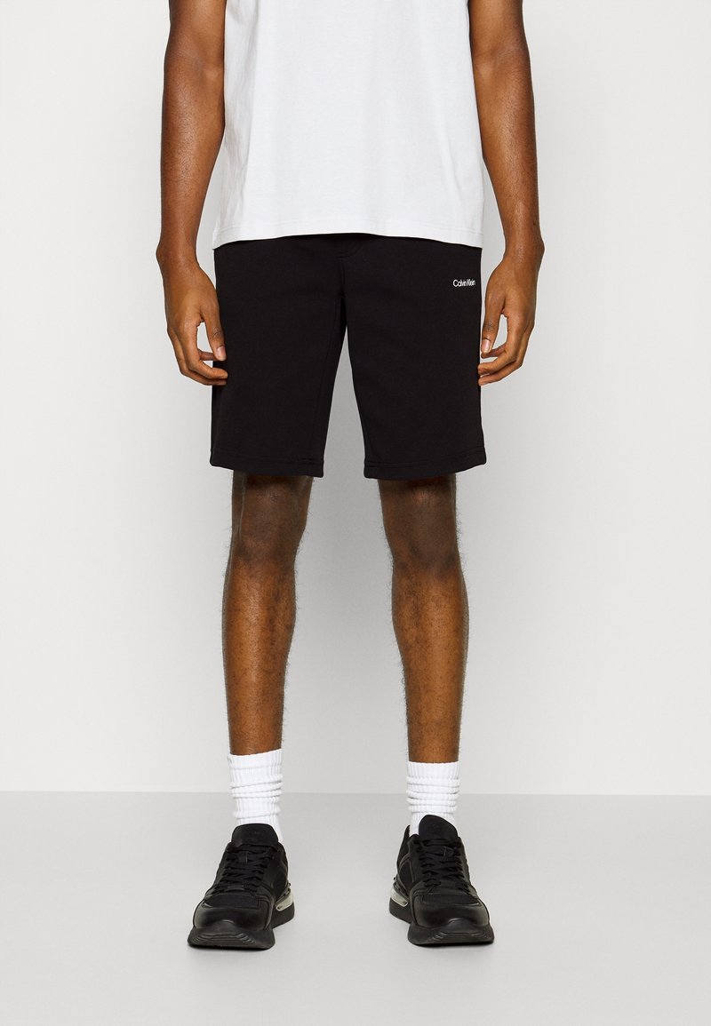 Homme portant un short Calvin Klein noir, des chaussettes blanches montantes, des baskets noires et un t-shirt blanc, debout devant un fond clair uni.