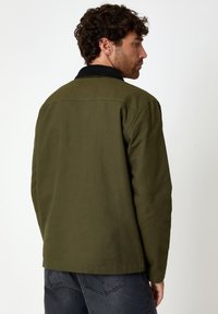 Giacca verde oliva con colletto in velluto a coste nero, texture semplice e vestibilità rilassata. Presenta un orlo dritto e dettagli di cucitura visibili.