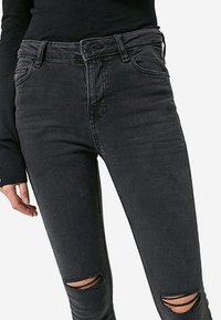 Černé džíny s vysokým pasem a slim fit, se dvěma roztrženými koleny, zapínáním na knoflík a předními kapsami. Vyrobeny z denimu s mírnou texturou.