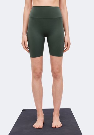 Personne debout pieds nus sur un tapis de yoga noir portant un short cycliste ajusté taille haute de couleur vert foncé sur un fond uni.