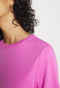 Fuchsiové bavlněné tričko s kulatým výstřihem a krátkými rukávy. Hladká textura s viditelným švem podél límce.