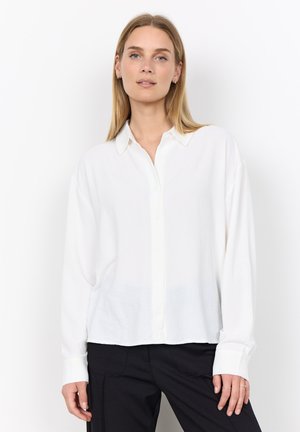 Young woman with straight blonde hair wearing a white button-up shirt and black pants standing against a white background.