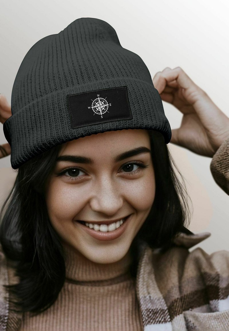 Black knit beanie featuring a compass design on a rectangular patch; textured fabric with a snug fit, worn slightly slouched.