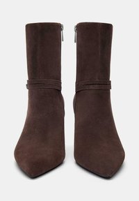 Stivali alla caviglia in suede marrone con punta appuntita, dotati di chiusura zip e due bande orizzontali attorno alla caviglia. Texture morbida e gambale alto.