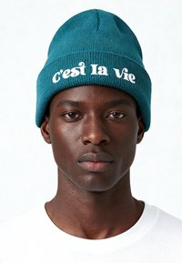 Cuffia lavorata a maglia color verde petrolio con la scritta "C'est la Vie" in bianco, caratterizzata da una vestibilità aderente e una texture a coste. Indossata sopra una maglietta bianca semplice.