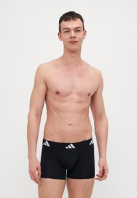 Svarta boxershorts med en vit märkesresår. Slät tygkvalitet, åtsittande design och shortsstil. Modellen står mot en enkel bakgrund.