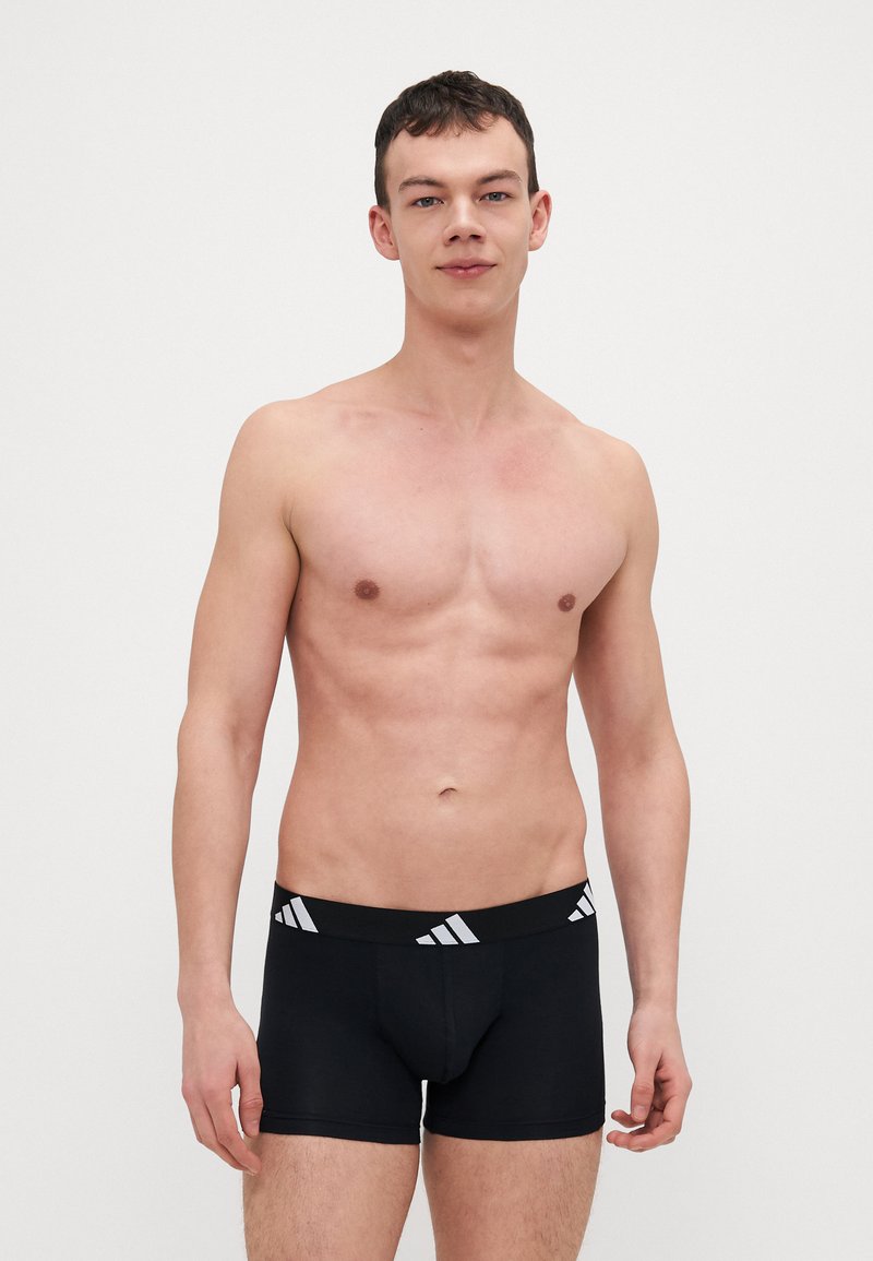 Svarta boxershorts med en vit märkesresår. Slät tygkvalitet, åtsittande design och shortsstil. Modellen står mot en enkel bakgrund.