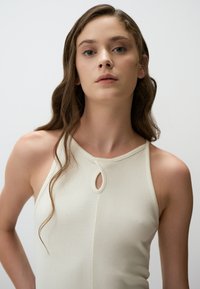 Top in cotone color crema a coste con un piccolo dettaglio a oblò sul colletto. Vestibilità slim con scollo a barca e spalline sottili. Texture liscia.