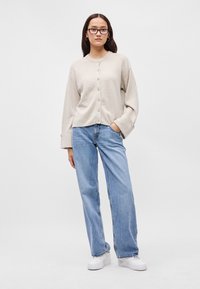 Μπεζ καπιτονέ cardigan με στρογγυλή λαιμόκοψη, χαλαρή εφαρμογή και μακριά μανίκια. Συνδυασμένο με ανοιχτό μπλε φαρδιών παντελονιών και λευκά αθλητικά παπούτσια.