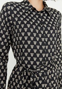 Černé šaty s květinovým potiskem, límečkovým výstřihm, dlouhými rukávy, knoflíkovým zapínáním vpředu a pásem ze stejného materiálu v pase. Hladká textura.