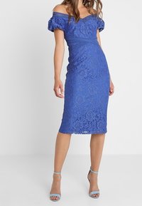 Žena ve světle modrých střevíčcích na kotník a modrých midi šatech s krajkou a odkrytými rameny, stojící na jednobarevném světlém pozadí.