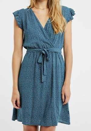 Kvinde iført en blå omslagskjole med korte ærmer, hvide polkaprikker og et bindebælte i taljen, stående mod en ensfarvet baggrund.