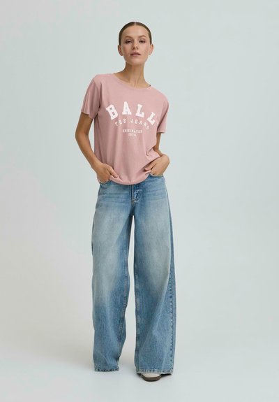 Žena oblečená v ružovom tričku s logom "BALL" a širokých modrých džínsach, stojaca s rukami v vreckách na svetlošedom pozadí.