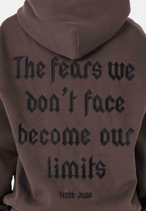 Bruine hoodie met tekst op de achterkant: "De angsten die we niet onder ogen zien, worden onze beperkingen" in zwart geborduurde gotische stijl lettertype.