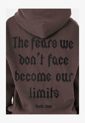 Brauner Hoodie mit Text auf dem Rücken: "Die Ängste, die wir nicht konfrontieren, werden zu unseren Grenzen" in schwarzem, gesticktem gotischem Schriftstil.