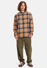 Flanellen shirt met een oranje, grijs en donkerbruin ruitpatroon, lange mouwen, knoopsluiting bij de kraag en een borstzak. Gecombineerd met groene broek.