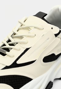 Zapatilla blanca y negra con parte superior de malla y cuero sintético, cordones planos, acentos texturizados y suela gruesa blanca.
