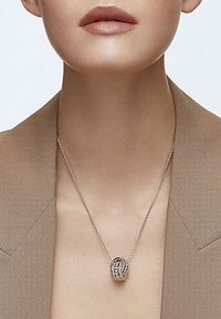 Collier à chaîne en argent avec un pendentif complexe et entrelacé orné de petites pierres précieuses, porté sur un blazer de couleur neutre.
