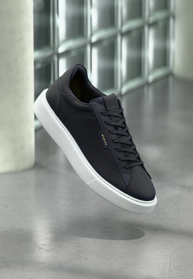 Marineblaue Ledersneaker mit einer weißen Gummisohle, runder Zehenform, flachen Schnürsenkeln und subtilen Branding-Details an der Seite. Strukturierte Oberfläche.