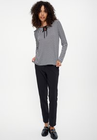Czarno-biały pasiasty top z długim rękawem i wiązaniem przy dekolcie, w połączeniu z czarnymi zwężanymi spodniami i błyszczącymi czarnymi loafersami.