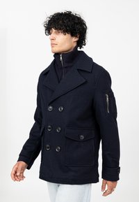 Granatowy wełniany płaszcz typu peacoat z podwójnym zapięciem, dużymi czarnymi guzikami, dwoma przednimi kieszeniami oraz kieszenią na ramieniu z suwakiem.