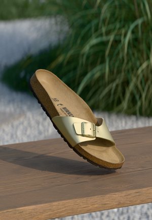 Złoty sandał Birkenstock z klamrą, leżący na drewnianej powierzchni na tle zielonych liści.
