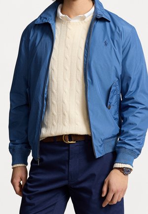Homme portant une veste à fermeture éclair bleue sur un pull crème à torsades, une chemise blanche à col, un pantalon marine, une ceinture marron et une montre-bracelet.