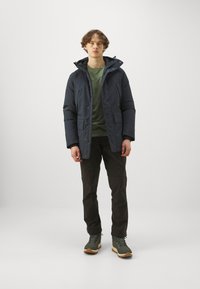 Navy puffer jacket with a hood, buttoned front, and two front pockets, worn over a green shirt and black pants, completed with green boots.