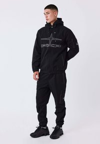Giovane in piedi, con una giacca nera con cappuccio e pantaloni neri abbinati con loghi, e sneaker nere su uno sfondo bianco.