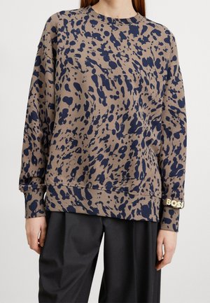 Femme portant un sweat-shirt beige avec des taches abstraites bleu foncé et un pantalon noir à jambes larges, avec l'inscription "BOSS" sur le poignet de la manche gauche.
