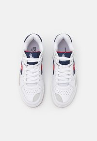 Pár bílých tenisek s námořnicky modrými a červenými akcenty, bílými tkaničkami a prošíváním, zobrazených shora na bílém pozadí.