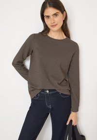 Brown long-sleeve top with a textured pattern, round neckline, and relaxed fit, paired with dark denim jeans displaying contrast stitching.