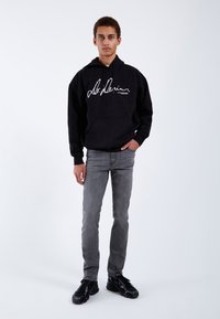 Sudadera negra con logo blanco escrito a mano, combinada con jeans gris ajustados y zapatillas negras. Diseño simple, textura suave, aspecto casual.