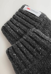 Grå stickade vantar med ribbad textur, med ljusa prickar och en 3M Thinsulate-etikett. Materialet verkar varmt och tjockt.