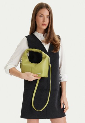 Donna che indossa un vestito nero senza maniche e una camicia bianca, che tiene una piccola borsa verde con tracolla lunga su sfondo neutro.