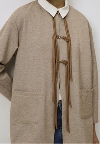 Beżowy, teksturowany płaszcz z białym kołnierzem, związanymi węzłami i srebrnymi akcentami. Dwie kieszenie z przodu, luźny krój i wzór w jodełkę.