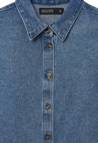 Camicia di denim blu chiaro, con colletto classico, bottoni marroni e tessuto testurizzato. L'etichetta recita "COLLEZIONE DENIM CALLIOPE", taglia S.
