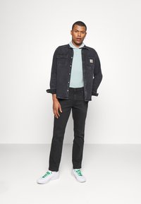 Chaqueta de mezclilla en gris oscuro con dos bolsillos en el pecho, camiseta polo azul claro, pantalones negros y zapatillas blancas con cordones verdes.