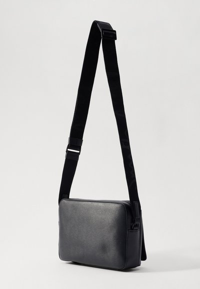 Borsa a tracolla rettangolare nera con esterno sintetico texture, tracolla regolabile e chiusura con zip. Dettaglio di branding discreto sulla tracolla.