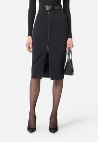 Jupe crayon noire ajustée avec un détail de fermeture éclair dorée à l'avant et une ceinture en simili cuir. Portée avec des collants noirs transparents et des talons hauts.